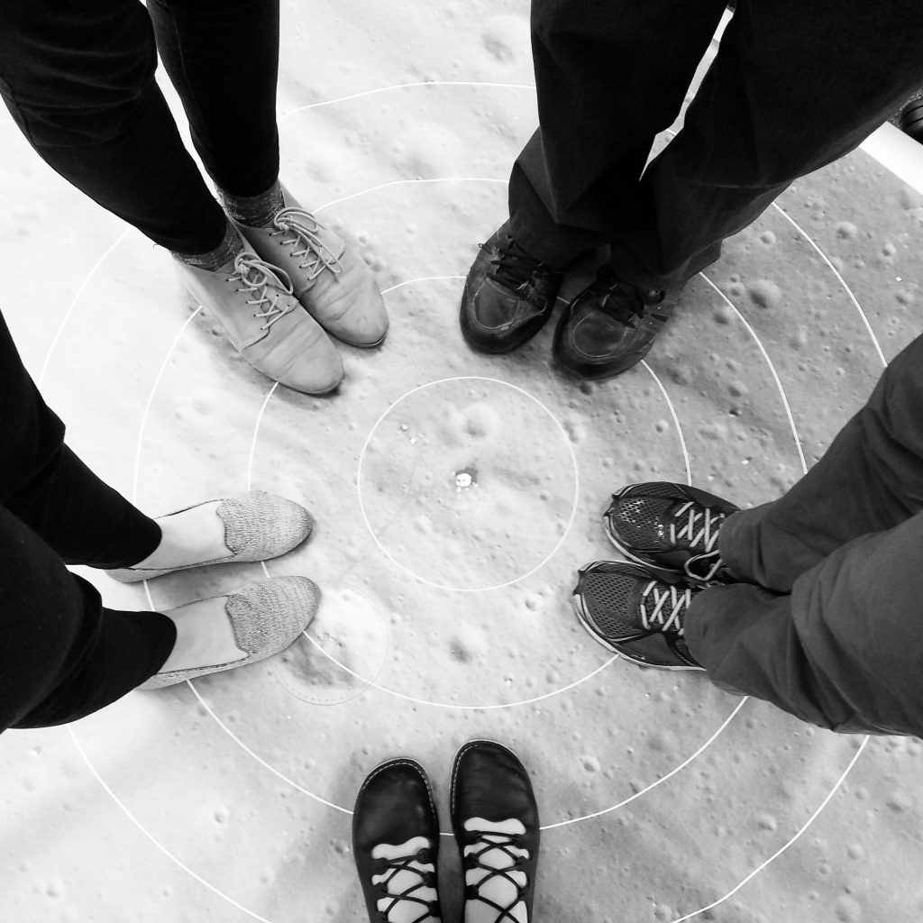 Black and white image showing 5 pairs of feet standing on a map of the lunar surface.