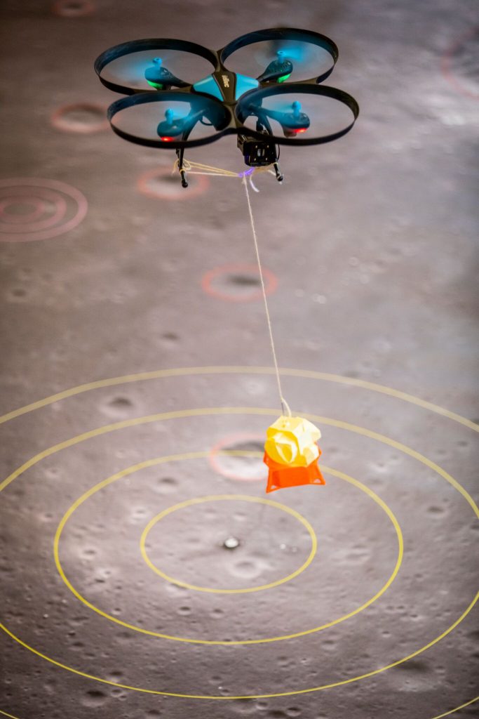 A flying drone carries a lunar lander above a map of the moon's surface. The landing spot is the actual site of the Apollo 11 landing. Other craters that teams will explore are circled in red.